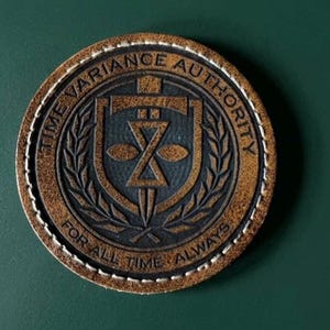 May include: A circular brown leather patch with white stitching along the edge. The center features a black embossed emblem with a shield, sword, and hourglass design. The text reads TIME VARIANCE AUTHORITY and FOR ALL TIME ALWAYS.