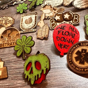 May include: A collection of wooden pins with various designs, including a green four-leaf clover, a red balloon with the text "We All Float Down Here", a green apple with a skull face, and a brown pin with a red star and the text "Boobs".