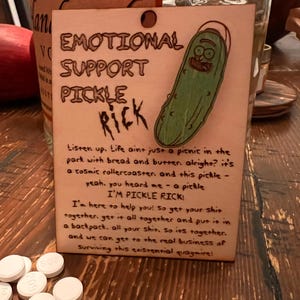 May include: A wooden sign with the text "EMOTIONAL SUPPORT PICKLE RICK" and a cartoon pickle illustration. The sign includes humorous text about life and getting things together. Several white pills are in the foreground.