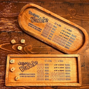 May include: Two wooden Farkle game boards with scoring charts. The boards are made of light brown wood and have black text. The text on the boards includes the names "Sheila's Farkle" and "Junars Farkle". The boards are on a wooden table.