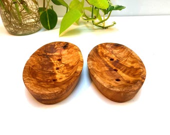May include: Two oval-shaped soap dishes carved from light and dark brown wood with visible grain patterns. Each dish features a smooth, concave surface with three small, circular drainage holes centered in the middle of the basin.