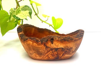 May include: A small, hand-carved wooden bowl made from olive wood, featuring a natural, irregular live edge with dark, textured bark detailing and a smooth, polished interior showcasing intricate, swirling brown wood grain patterns.