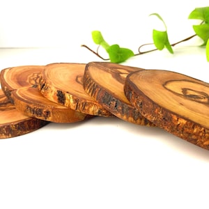 Peut inclure: Un ensemble de quatre sous-verres circulaires en bois avec des bords en écorce naturelle. Chaque sous-verre présente un motif de grain de bois brun clair et foncé distinct avec des anneaux visibles, soulignant la texture organique.