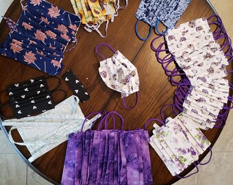 May include: A variety of fabric face masks in different colors and patterns, including purple, blue, white, and black. The masks are arranged on a wooden table.