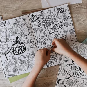 May include: A set of five coloring books with Halloween and fall themes. The books feature illustrations of ghosts, pumpkins, candy, and other spooky and festive imagery. The books are open and a child's hands are coloring one of the pages.