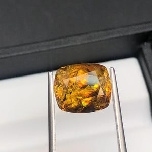 Pode incluir: Uma gema facetada, com corte almofada, segurada por pin&ccedil;as. A gema exibe uma vibrante variedade de cores, incluindo dourado, laranja e toques de verde. As facetas da pedra refletem a luz, criando um efeito cintilante. O fundo &eacute; cinza escuro.