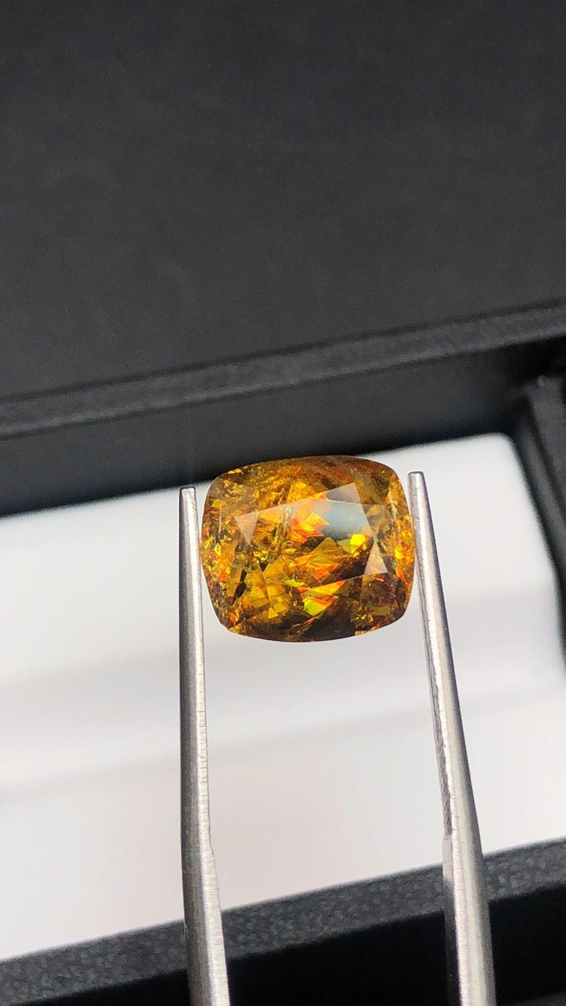 Pode incluir: Uma gema facetada, com corte almofada, segurada por pin&ccedil;as. A gema exibe uma vibrante variedade de cores, incluindo dourado, laranja e toques de verde. As facetas da pedra refletem a luz, criando um efeito cintilante. O fundo &eacute; cinza escuro.