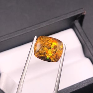 Pode incluir: Uma pedra preciosa de corte quadrado com um interior vibrante e multicolorido, segurada por pin&ccedil;as de prata. A gema exibe tons de ouro, laranja e verde, com uma superf&iacute;cie polida. A pedra preciosa est&aacute; colocada contra uma caixa preta com interior branco.