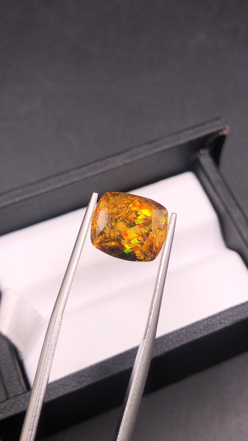 Pode incluir: Uma pedra preciosa de corte quadrado com um interior vibrante e multicolorido, segurada por pin&ccedil;as de prata. A gema exibe tons de ouro, laranja e verde, com uma superf&iacute;cie polida. A pedra preciosa est&aacute; colocada contra uma caixa preta com interior branco.