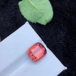 May include: A pink, square-shaped gemstone with a cushion cut. The gemstone is on a white surface.