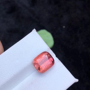 May include: A pink, rectangular gemstone with a faceted cut. The gemstone is on a white surface.