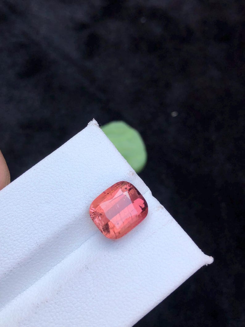 May include: A pink, rectangular gemstone with a faceted cut. The gemstone is on a white surface.