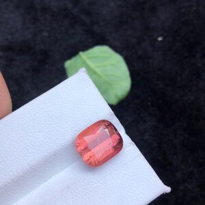 May include: A pink, square-shaped gemstone with a cushion cut. The gemstone is on a white surface.
