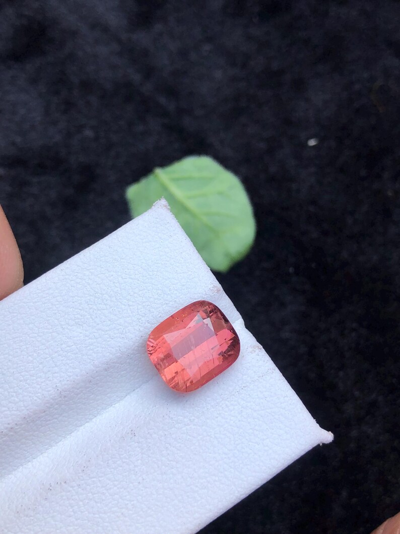 May include: A pink, square-shaped gemstone with a cushion cut. The gemstone is on a white surface.