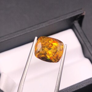 Pode incluir: Uma pedra preciosa de corte almofada presa por pin&ccedil;as. A gema tem uma cor amarelo-dourado quente com flashes internos de laranja, vermelho e verde. A pedra est&aacute; sobre um fundo preto, sugerindo uma apresenta&ccedil;&atilde;o.