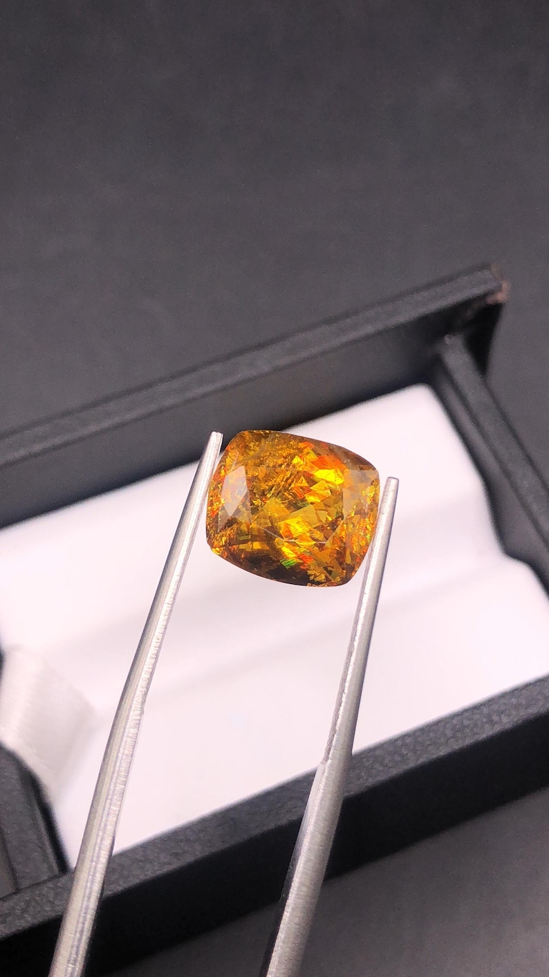 Pode incluir: Uma pedra preciosa de corte almofada presa por pin&ccedil;as. A gema tem uma cor amarelo-dourado quente com flashes internos de laranja, vermelho e verde. A pedra est&aacute; sobre um fundo preto, sugerindo uma apresenta&ccedil;&atilde;o.