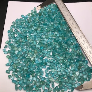 May include: A large quantity of small, teal blue, faceted gemstones. The gemstones are spread out on a white surface with a ruler for scale.