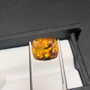 Pode incluir: Uma pedra preciosa de corte almofada segurada por pin&ccedil;as. A gema apresenta uma exibi&ccedil;&atilde;o vibrante de cores, incluindo laranja, amarelo e toques de verde. A pedra &eacute; colocada sobre um fundo preto, real&ccedil;ando seu brilho. As pin&ccedil;as s&atilde;o prateadas.