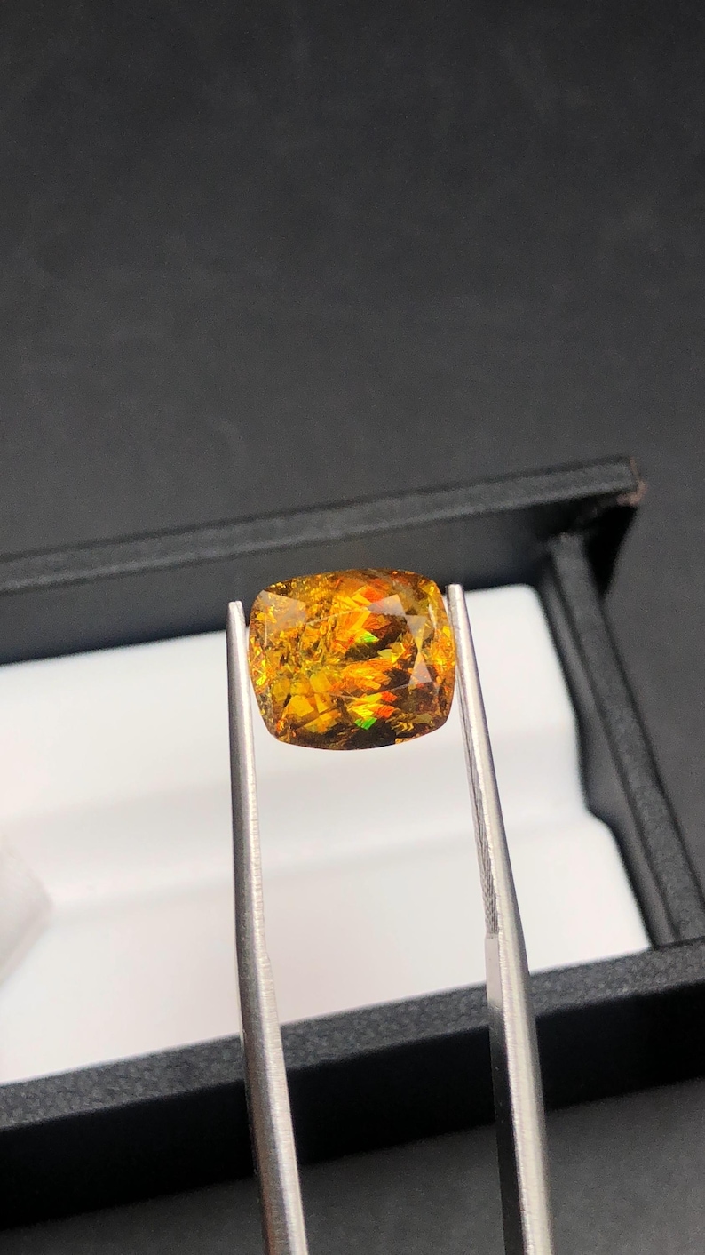 Pode incluir: Uma pedra preciosa de corte almofada segurada por pin&ccedil;as. A gema apresenta uma exibi&ccedil;&atilde;o vibrante de cores, incluindo laranja, amarelo e toques de verde. A pedra &eacute; colocada sobre um fundo preto, real&ccedil;ando seu brilho. As pin&ccedil;as s&atilde;o prateadas.
