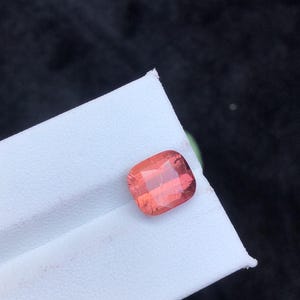May include: A pink gemstone with a cushion cut. The gemstone is on a white surface.