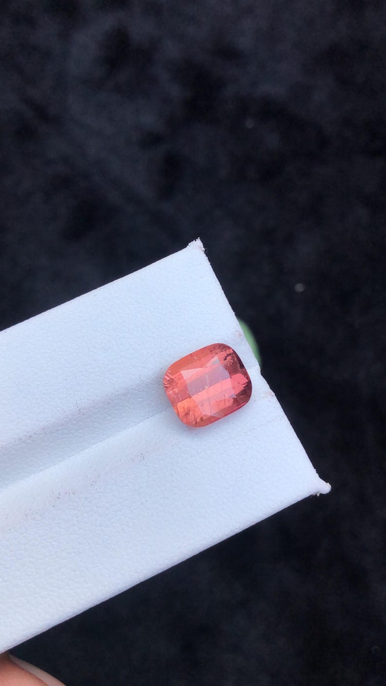 May include: A pink gemstone with a cushion cut. The gemstone is on a white surface.