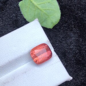 May include: A square-shaped, reddish-pink gemstone with a slight shimmer. The gemstone is on a white surface.