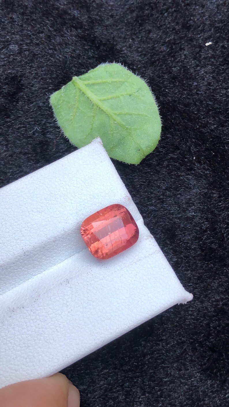 May include: A square-shaped, reddish-pink gemstone with a slight shimmer. The gemstone is on a white surface.