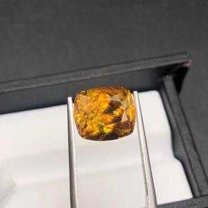 Pode incluir: Uma pedra preciosa facetada, de corte quadrado, segurada por pin&ccedil;as. A gema tem um tom quente de laranja dourado com flashes verdes e vermelhos. &Eacute; apresentada contra um fundo preto, sugerindo uma exibi&ccedil;&atilde;o de joias ou pedras preciosas.