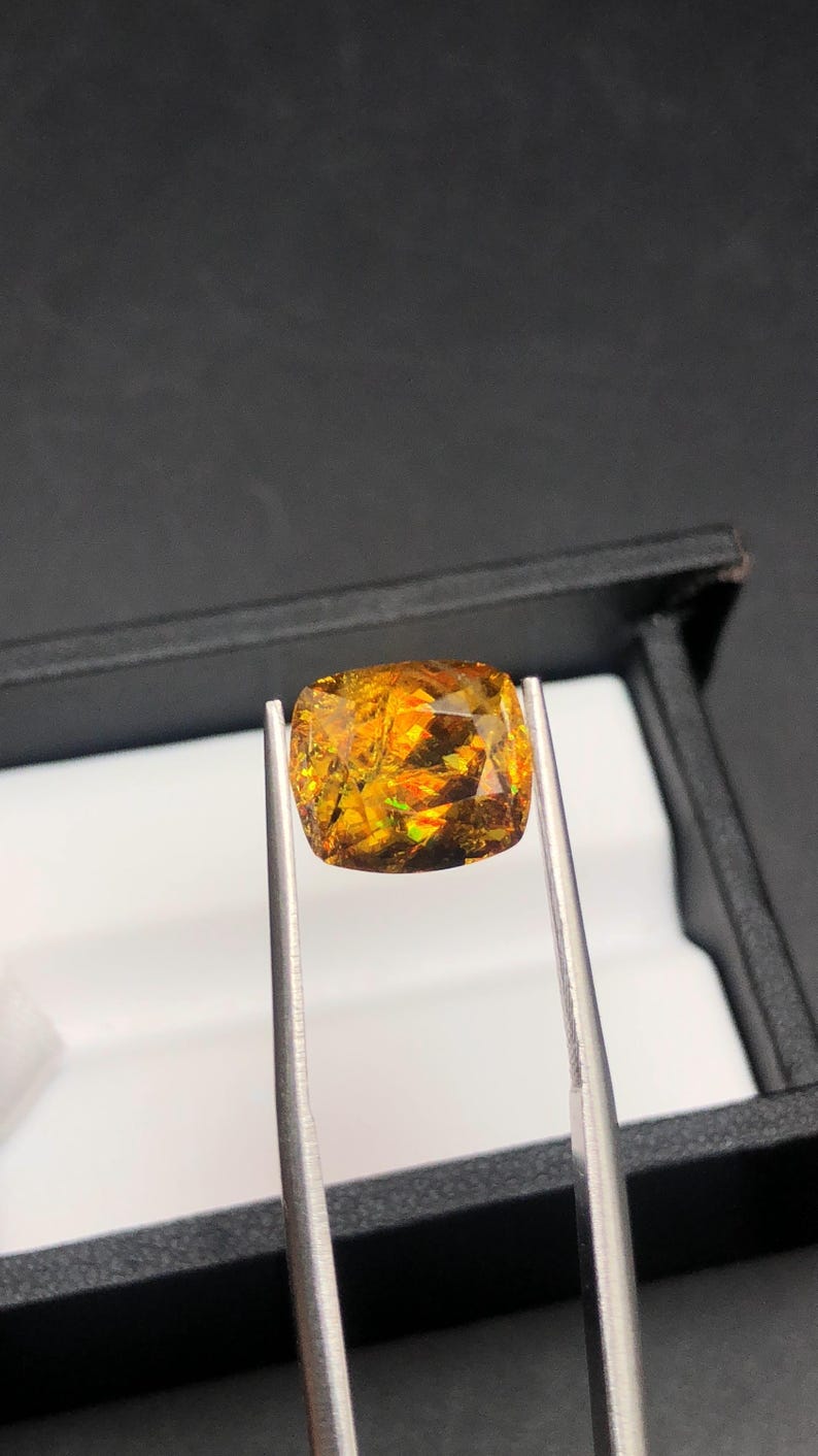 Pode incluir: Uma pedra preciosa facetada, de corte quadrado, segurada por pin&ccedil;as. A gema tem um tom quente de laranja dourado com flashes verdes e vermelhos. &Eacute; apresentada contra um fundo preto, sugerindo uma exibi&ccedil;&atilde;o de joias ou pedras preciosas.