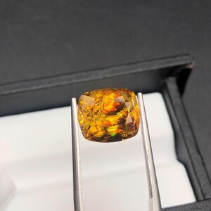Pode incluir: Uma gema facetada, com corte almofada, segurada por pin&ccedil;as. A gema exibe uma vibrante variedade de cores, incluindo laranja, amarelo e toques de verde. Est&aacute; colocada sobre um fundo escuro, real&ccedil;ando o seu brilho e clareza.