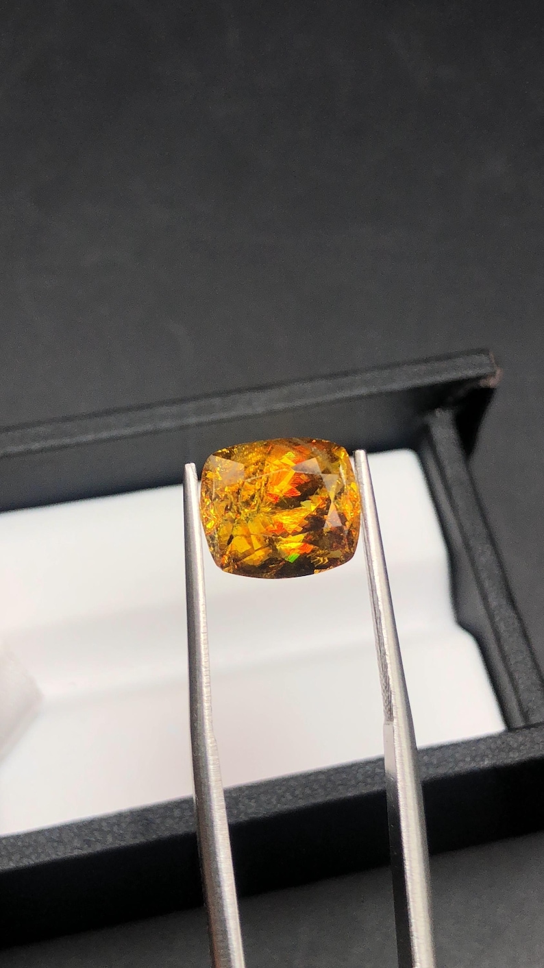 Pode incluir: Uma gema facetada, com corte almofada, segurada por pin&ccedil;as. A gema exibe uma vibrante variedade de cores, incluindo laranja, amarelo e toques de verde. Est&aacute; colocada sobre um fundo escuro, real&ccedil;ando o seu brilho e clareza.