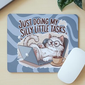 Pode incluir: Tapete de rato retangular com um gato de desenho animado com óculos usando um laptop. O texto "JUST DOING MY SILLY LITTLE TASKS" está presente. Uma xícara de café, um rato branco e lápis também são visíveis.