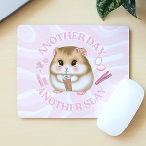 May include: A pink mouse mat featuring a cartoon hamster with a drink, and makeup illustrations. The text "ANOTHER DAY ANOTHER SLAY" is printed on the mat. A white computer mouse is positioned beside the mat.