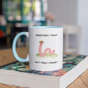 May include: White ceramic mug with a light blue interior and handle. The mug features a cartoon worm wearing a cowboy hat, with the text "SOMETIMES I THINK BUT THEN I FORGET". The mug is sitting on a book.