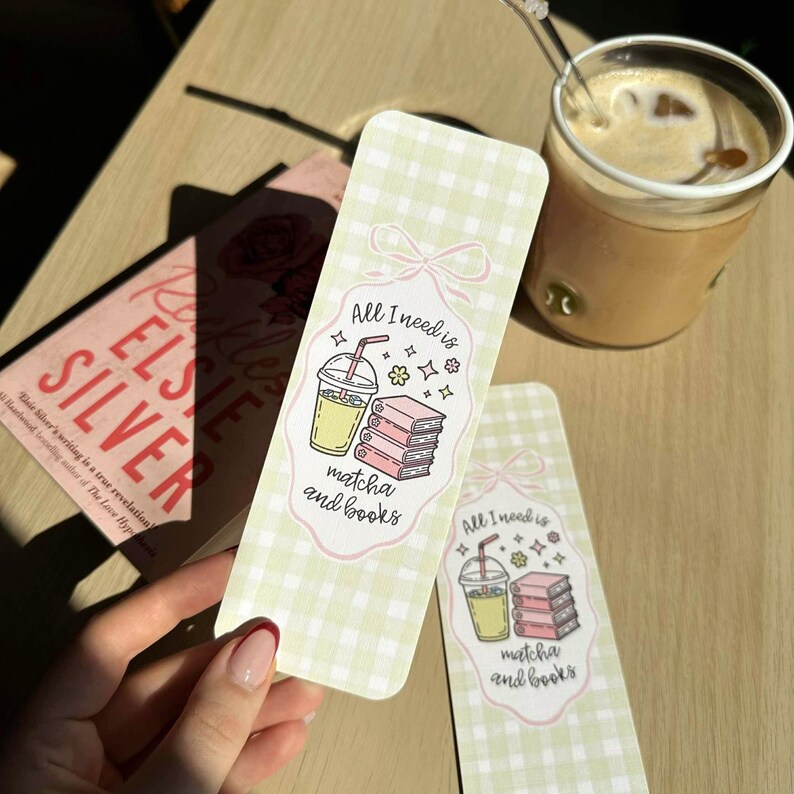 Op de afbeelding: Twee bladwijzers met de tekst "All I need is matcha and books" en een illustratie van matcha-drank en boeken. De bladwijzers hebben een lichtgroen ruitpatroon en een roze strikdesign. Op de achtergrond een boek met de titel "Elsie Silver".