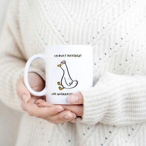 Puede incluir: Taza de cer&aacute;mica blanca con un dibujo de un ganso con sombrero y el texto "I'M BUILT DIFFERENT LIKE INCORRECT". La taza tiene un asa curva y la sostiene una persona que lleva un jersey color crema.
