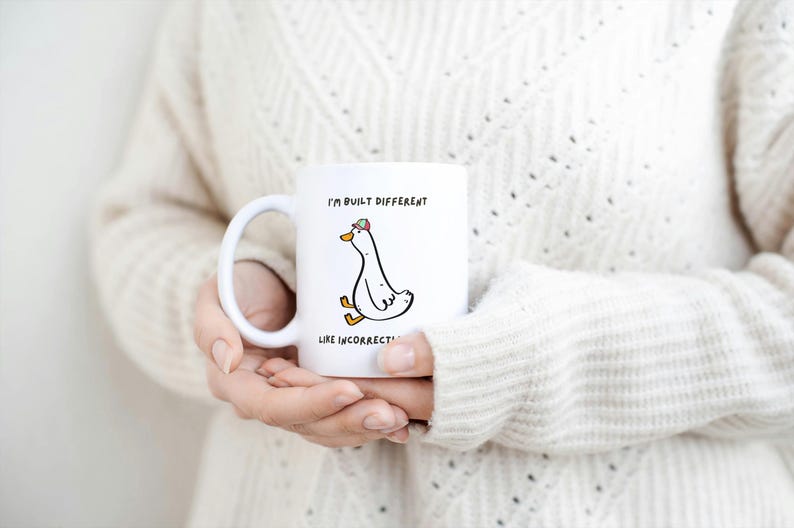 Puede incluir: Taza de cer&aacute;mica blanca con un dibujo de un ganso con sombrero y el texto "I'M BUILT DIFFERENT LIKE INCORRECT". La taza tiene un asa curva y la sostiene una persona que lleva un jersey color crema.