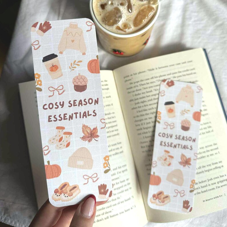 May include: Two rectangular bookmarks with a cozy season theme. The bookmarks feature illustrations of a sweater, coffee cup, pumpkin, and other fall-themed items. The text "COSY SEASON ESSENTIALS" is printed on the bookmarks. One bookmark is in a book, and the other is held by a person.