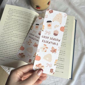May include: Two rectangular bookmarks with a fall theme, featuring pumpkins, leaves, and cozy items. The text "COSY SEASON ESSENTIALS" is printed on the bookmarks. The bookmarks are held over an open book.