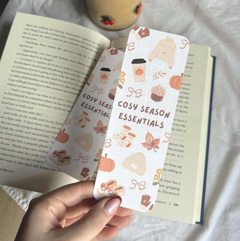 May include: Two rectangular bookmarks with a fall theme, featuring pumpkins, leaves, and cozy items. The text "COSY SEASON ESSENTIALS" is printed on the bookmarks. The bookmarks are held over an open book.