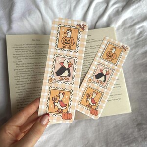 Op de afbeelding: Twee decoratieve bladwijzers met een Halloween-thema. Elke bladwijzer heeft drie postzegelachtige illustraties van een eend met een pompoen, een pingu&iuml;n in een cape en een eend met een drietand. De bladwijzers hebben een lichtoranje en wit geruit patroon.
