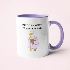 May include: White ceramic mug with a purple handle and rim. The mug features a cartoon duck wearing a pink robe and holding a small bowl. Text on the mug reads: "BESTIE, I'M ABOUT TO CRASH TF OUT."