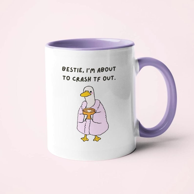 May include: White ceramic mug with a purple handle and rim. The mug features a cartoon duck wearing a pink robe and holding a small bowl. Text on the mug reads: "BESTIE, I'M ABOUT TO CRASH TF OUT."