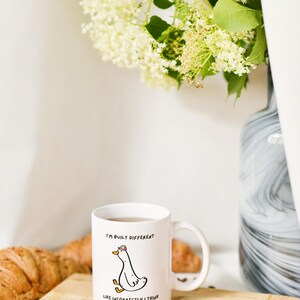 Puede incluir: Taza de cer&aacute;mica blanca con una ilustraci&oacute;n de un ganso de dibujos animados y el texto "I'M BUILT DIFFERENT LIKE INCORRECTLY I THINK." La taza est&aacute; sobre una tabla de madera con un cruas&aacute;n y una cuchara. Un jarr&oacute;n con flores est&aacute; en el fondo.