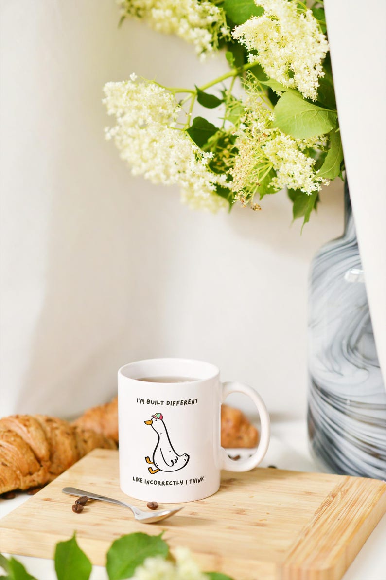 Puede incluir: Taza de cer&aacute;mica blanca con una ilustraci&oacute;n de un ganso de dibujos animados y el texto "I'M BUILT DIFFERENT LIKE INCORRECTLY I THINK." La taza est&aacute; sobre una tabla de madera con un cruas&aacute;n y una cuchara. Un jarr&oacute;n con flores est&aacute; en el fondo.
