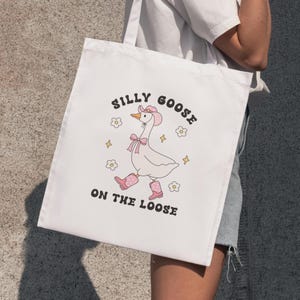 May include: White tote bag with a cartoon goose wearing a pink cowboy hat and boots. The text on the bag reads "Silly Goose on the Loose".