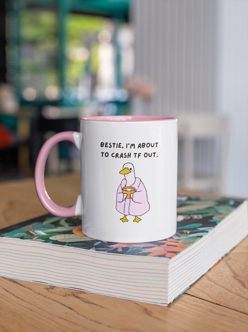 May include: White ceramic mug with a pink interior and handle. The mug features a cartoon duck wearing a pink robe and holding a cup, with the text "BESTIE, I'M ABOUT TO CRASH TF OUT."