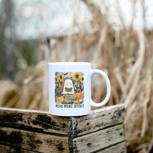 May include: White ceramic mug featuring a Halloween-themed design. The design includes a ghost reading a book, sunflowers, pumpkins, and the text "READ MORE BOOKS". The mug is set against a blurred outdoor background.