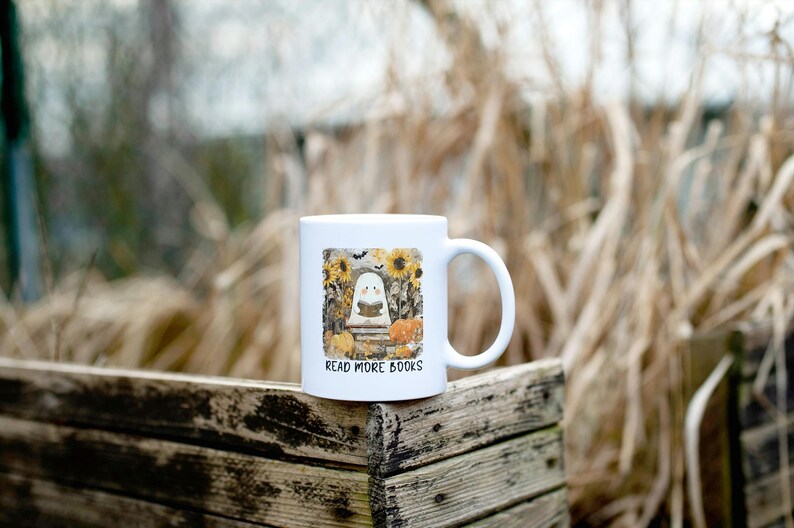 May include: White ceramic mug featuring a Halloween-themed design. The design includes a ghost reading a book, sunflowers, pumpkins, and the text "READ MORE BOOKS". The mug is set against a blurred outdoor background.