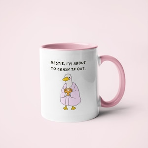 May include: White ceramic mug with a pink interior and handle. The mug features a cartoon duck wearing a pink robe and holding a golden cup. Text on the mug reads, "BESTIE, I'M ABOUT TO CRASH TF OUT."